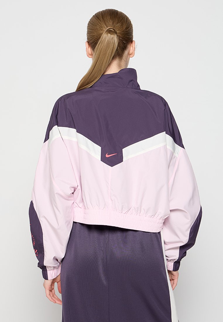 Mujer con coleta rubia que lleva una chaqueta cortavientos de Nike en púrpura y rosa, vista desde la espalda contra un fondo liso.