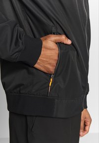 Sort bomberjakke med en glat tekstur, der har en lynlåslomme og elastiske manchetter. Inkluderer orange hardware-detaljer på lynlåsen.