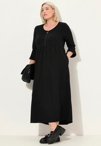 Schwarzes Langarmkleid mit runder Ausschnitt und Reißverschlusdetail. Hat ein tailliertes Oberteil und einen ausgestellt Rock mit seitlichen Taschen.