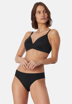 Mujer con sujetador negro sin costuras y ropa interior a juego de corte alto, de pie con las manos entrelazadas y mirando hacia un lado sobre un fondo blanco.