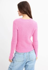 Mujer con cabello largo castaño que lleva un suéter de lana rosa y pantalones cargo de mezclilla azul claro, de lado frente a un fondo blanco.