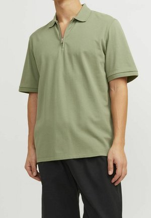 T-shirt polo à manches courtes en vert olive, fabriqué en tissu texturé. Présente un col zippé à demi et une coupe décontractée. Accompagné de pantalons noirs.