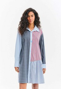 Žena s dlouhými kudrnatými vlasy, která má na sobě dlouhé šaty s nášivkami a kostkovaným vzorem v modrých, červených a černých tónech, na bílém pozadí.