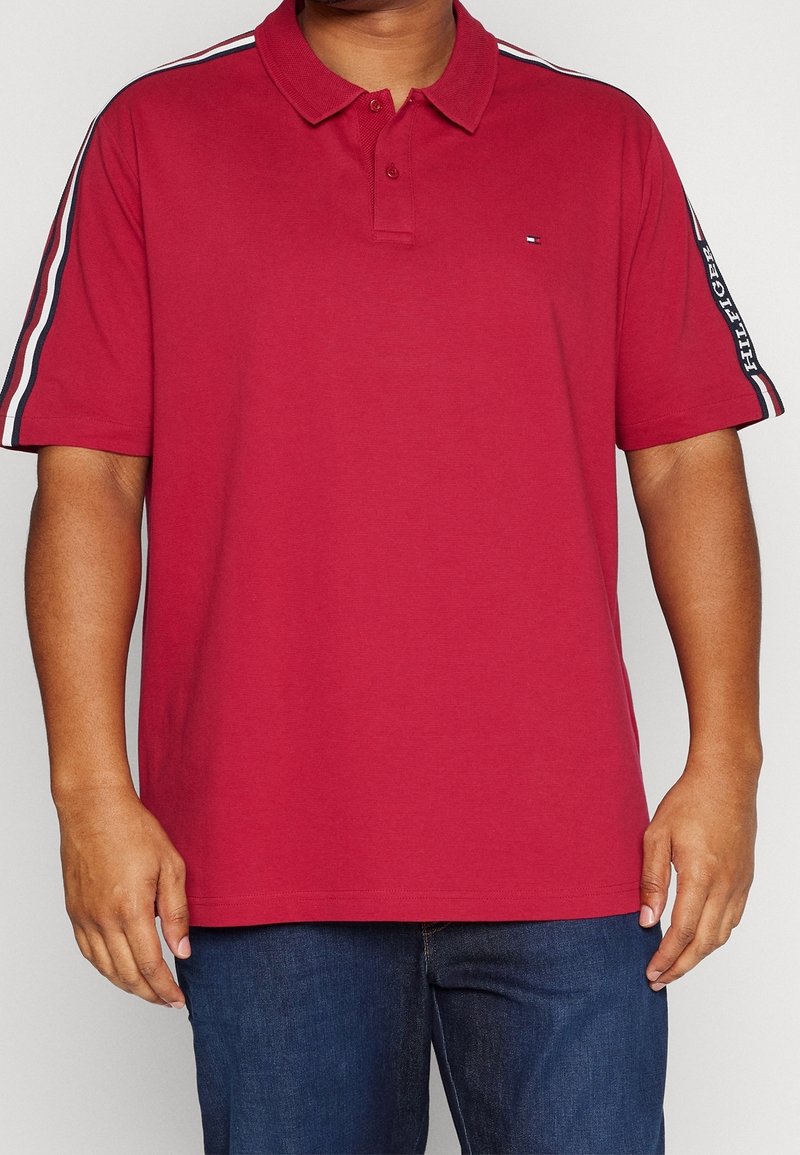 Homme portant un polo rouge à manches courtes avec des rayures blanches et noires sur les manches et un petit logo sur la poitrine gauche, associé à un jean bleu.