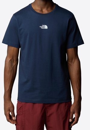 Homme portant un t-shirt bleu marine à manches courtes avec le logo blanc The North Face et un short cargo rouge, debout devant un fond blanc.
