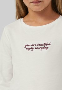 Valge pikkade varrukatega särk ümmarguse kaelusega, millel on bordoopunase värviga tikitud tekst "sa oled ilus, naudi iga päeva". Pehme puuvillane tekstuur.