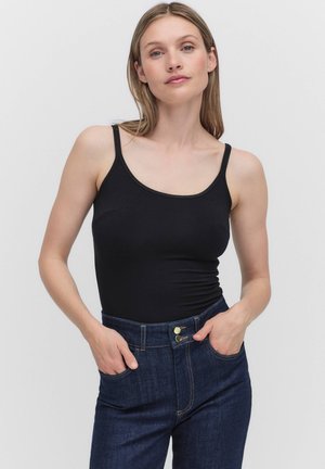 Kvinde med lyst hår iført en tætsiddende sort ærmeløs top og højtsiddende blå jeans, stående med hænderne i forlommerne.
