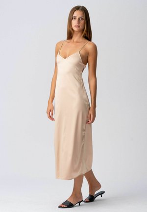 Žena stojaca v bezových saténových midi šatách bez rukávov s tenkými ramienkami a čiernymi otvorenými sandálmi s nízkym podpätkom na jednofarebnom pozadí.