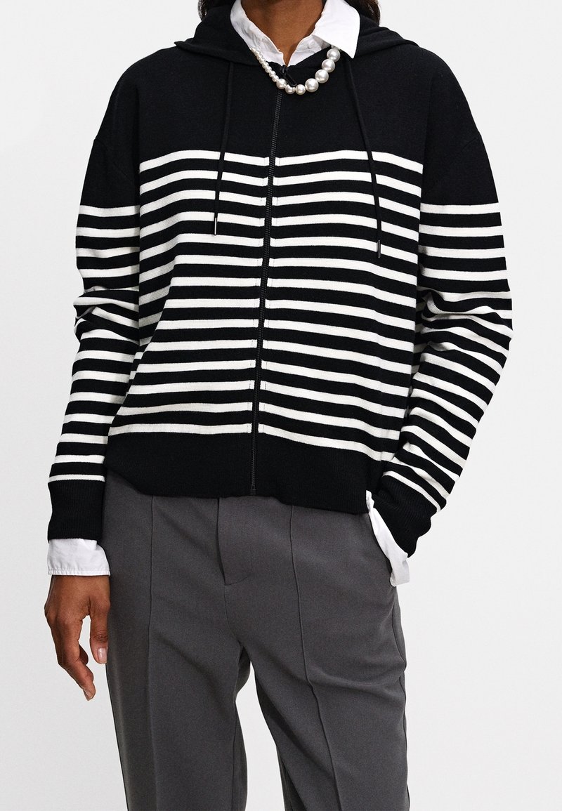 Personne portant un sweat à capuche zippé à rayures horizontales noires et blanches sur une chemise blanche à col et un pantalon gris, avec un collier de perles.