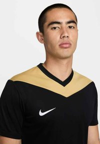 Junger Mann mit kurzen Haaren, der ein schwarzes und goldenes Nike-Sportshirt trägt und mit neutralem Ausdruck direkt nach vorne schaut.