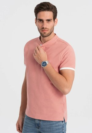 Camisa polo de manga corta, ligera, de color rosa con cuello mandarín y acentos blancos en las mangas, combinada con un reloj plateado y pantalones vaqueros.