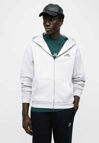 Άνδρας που φοράει λευκή ζακέτα με φερμουάρ πάνω από πράσινο πουκάμισο, μαύρο παντελόνι και μαύρο δερμάτινο καπέλο μπέιζμπολ, στέκεται με το ένα χέρι στην τσέπη.