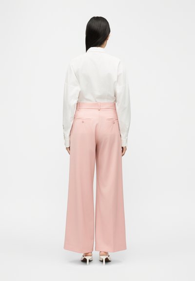 Γυναίκα με μακριά σκούρα μαλλιά που φοράει λευκό πουκάμισο με μακριά μανίκια, ροζ παντελόνια ψηλής μέσης με φαρδιά πόδια και λευκά παπούτσια με τακούνι, γυρισμένη προς τα πίσω.
