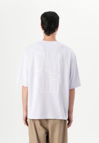 Fialové bavlněné tričko s velkými bílými písmeny, které na zádech hlásají "CALVIN KLEIN". Krátké rukávy, volný střih, minimalistický design.