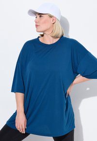 T-shirt blu grande con maniche corte, realizzato in un tessuto liscio, abbinato a un cappellino bianco. Vestibilità ampia e scollatura tonda. Leggings neri visibili.