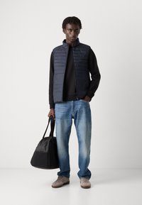 Colete acolchoado azul-marinho sobre um suéter preto, combinado com jeans azul claro e ténis bege. Segura uma mala de viagem preta. Design simples e moderno.