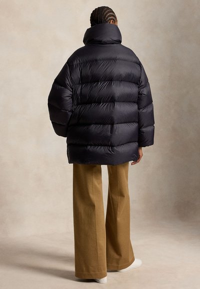 Person med fletninger iført en sort oversized dunkappe, brede brune bukser og hvide sko, set bagfra mod en neutral baggrund.