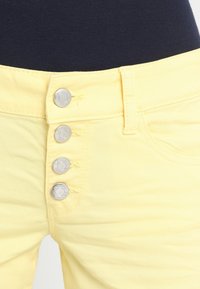 Los shorts de mezclilla amarillos presentan una cintura alta con cuatro botones de presión plateados y detalles de costura sutiles. Tienen una textura suave en toda su superficie.