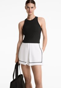 Svart ribbad linne utan ärmar, i kombination med vita shorts med svarta pipingsdetaljer. Modellen håller en svart tote-väska.