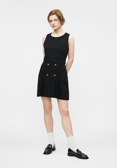 Νεαρό άτομο που φοράει μαύρο αμάνικο φόρεμα με χρυσά κουμπιά, λευκές κάλτσες και μαύρα μοκασίνια, στέκεται μπροστά σε απλό λευκό φόντο.