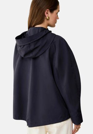 Donna con capelli castani che indossa orecchini d'oro, una giacca con cappuccio blu navy e pantaloni bianchi, vista di spalle su uno sfondo semplice.