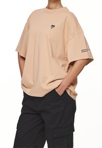 Beige oversized tričko z měkké bavlny, s malým černým logem na hrudi a textem na rukávu. Krátké rukávy s volným střihem.