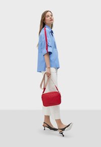 Borsa a mano rettangolare rossa con logo in rilievo, abbinata a una camicia azzurra chiara con dettagli a strisce rosse e pantaloni bianchi a vita alta. Scarpe slingback nere.