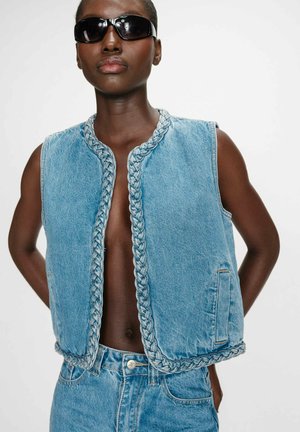 Personne à la peau foncée portant des lunettes de soleil noires, un gilet en denim bleu sans manches avec une bordure tressée, et un jean taille haute en denim bleu, debout les mains derrière le dos.