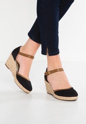Zwarte suède espadrille sleehakken met een beige enkelriem, voorzien van geweven jute zolen. De schoen heeft een ronde neus en een textuur oppervlak.