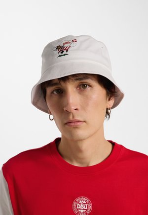 Jonge man draagt een witte bucket hat met een sportcartoon en de Deense vlag, en een rood shirt met het logo van de Deense voetbalbond.