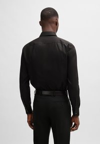 Camisa negra de manga larga con una textura suave, cuello clásico y puños con botones, combinada con pantalones negros elegantes y un cinturón a juego.