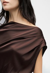 Γυναίκα με σκούρα μαλλιά που φοράει λαμπερό καφέ σατέν τοπ με ακάλυπτους ώμους πάνω σε απλό ανοιχτό φόντο.