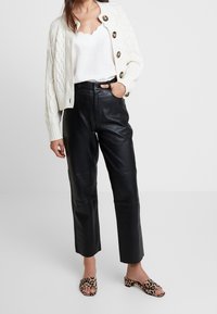 Weißer, kabelgenähter, verkürzter cardigan mit großen Knöpfen, kombiniert mit schwarzen, hochtaillierten Lederhosen und offenen Mules mit Leopardenmuster.