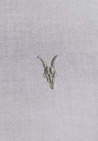 Tessuto grigio con una piccola e intricata teschio verde ricamato con corna curve, impostato su uno sfondo testurizzato.