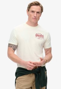 Cremefarvet T-shirt med korte ærmer og rødt grafisk logo, båret med en grøn sweater knyttet om taljen. Let og blød stof, afslappet design.
