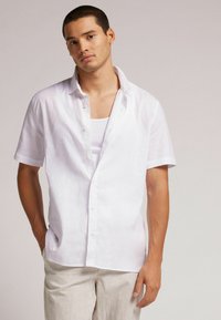Camisa de lino blanca de manga corta con tapeta de botones, cuello relajado y un corte ligeramente holgado, combinada con pantalones de lino de color claro.