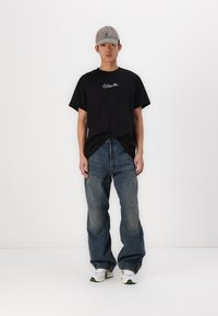 T-shirt en coton noir avec un logo blanc, associé à un jean en denim bleu au détail de patchwork et à la coupe décontractée. Porté avec une casquette grise et des baskets.