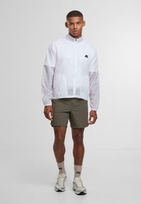 Mandlig model iført hvid lynlåsjakke, olivengrønne shorts, beige sokker og grå sneakers, der står mod en ensfarvet lys baggrund.