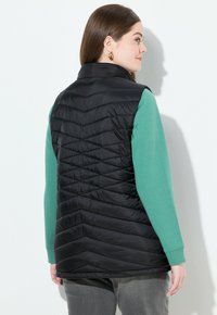 Schwarze gesteppte Weste mit hohem Kragen und einem Zickzackmuster. Getragen über einem grünen Langarmhemd, kombiniert mit grauen Hosen.