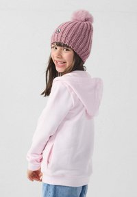 Felpa con cappuccio di colore rosa chiaro, dotata di tasche laterali. Abbinata a un berretto di maglia rosa a coste con un pon pon sulla sommità e una patch con logo.