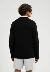 Czarny sweter prążkowany z okrągłym dekoltem i długimi rękawami, z teksturowanym wzorem dzianiny. Nosi się go na jasnoszarych szortach.