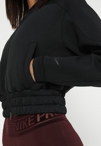 Frau trägt eine schwarze, cropped Jacke mit Reißverschluss und elastischem Bund, Hand in der Tasche, kombiniert mit dunkelroten Nike Pro Leggings.