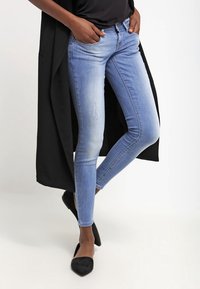 Jeans ajustados de un azul claro con detalles desgastados, combinados con un cárdigan largo negro y tacones de punta negra, creando una silueta estilizada.