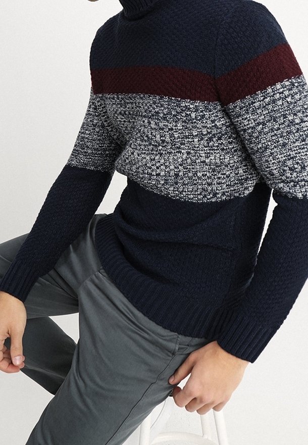Homme portant un pull en maille rayé bleu marine, gris et bordeaux avec un pantalon gris, assis sur un tabouret blanc devant un fond uni.