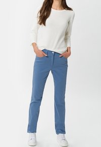 Celana katun biru muda dengan potongan slim, saku depan, dan penutupan kancing, dipadukan dengan atasan lengan panjang bergaris putih dan sepatu sneakers putih.