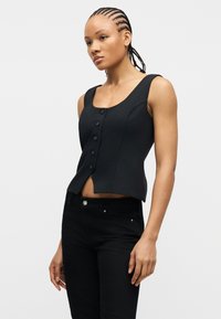 Γυναίκα με πλεξούδες, που φοράει μαύρο αμάνικο τοπ με κουμπιά και μαύρο παντελόνι, στέκεται μπροστά σε ένα απλό φωτεινό φόντο.