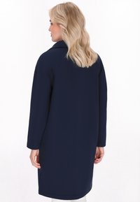 Tamnoplavi kaput s dugim rukavima, ravnog kroja, ovratnikom i bočnim džepovima. Glatka tkanina i minimalni detalji šivanja. Prikazan stražnji pogled.