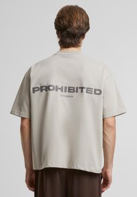 Lys grå T-skjorte med korte ermer som har fet svart tekst "PROHIBERT" og "10119, BERLIN" på baksiden; glatt, uformell passform med avslappet design.