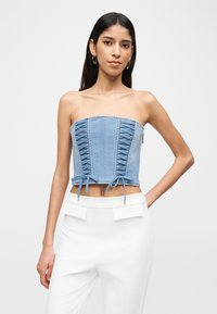 Γυναίκα που φοράει κορσέ τοπ από ανοιχτό μπλε denim χωρίς τιράντες με λεπτομέρειες δέσιμο και άσπρο ψηλόμεσο παντελόνι με καλύμματα τσεπών.