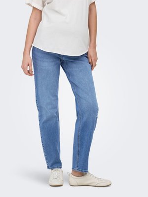 Persona che indossa jeans azzurri a gamba dritta, top bianco a maniche corte e sneakers bianche con lacci, in piedi davanti a uno sfondo semplice.